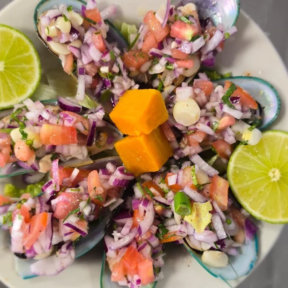 Mussels topped with tomato, red onion, herbs, and orange garnish, served with lime halves on a white plate.