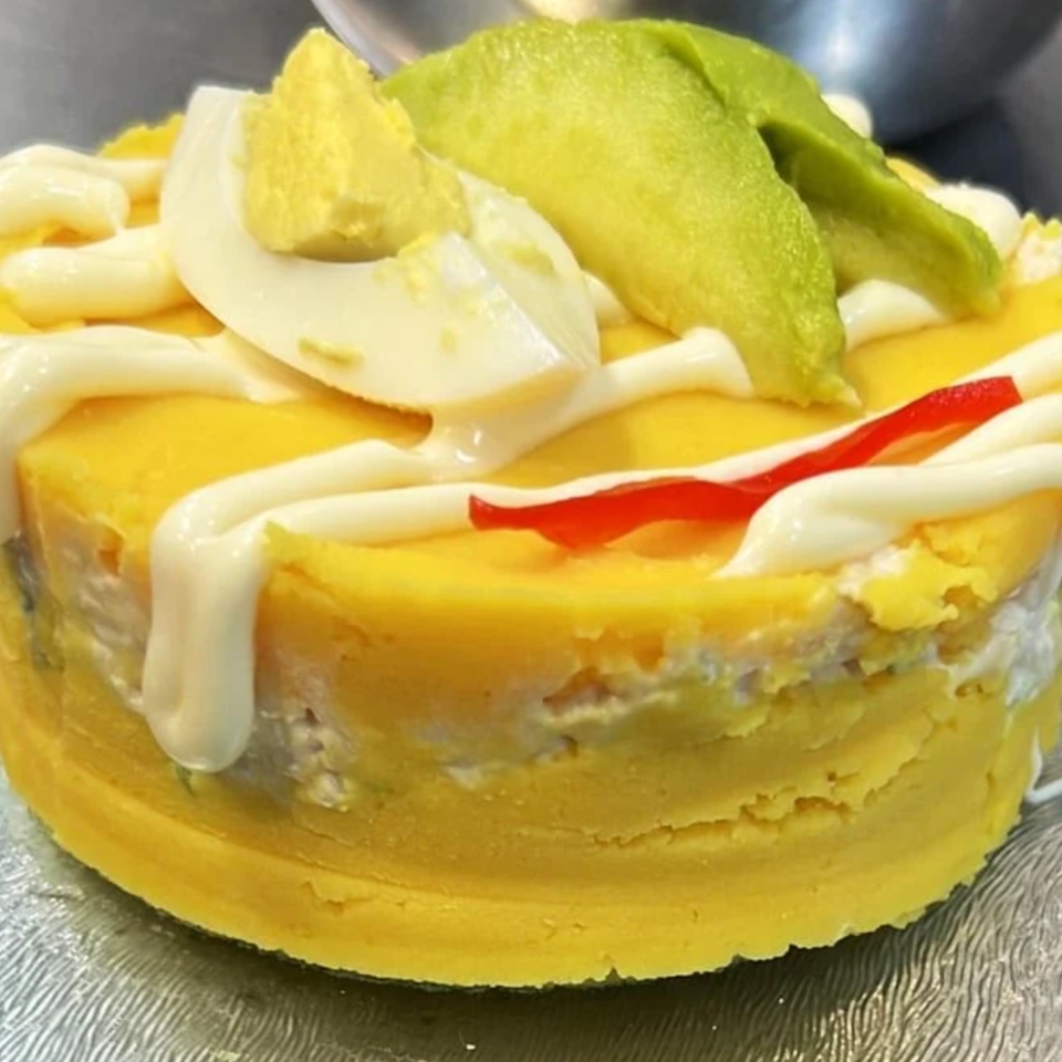 Close-up of a yellow savory pastry topped with mayonnaise, sliced avocado, onion, and red pepper on a metal tray.